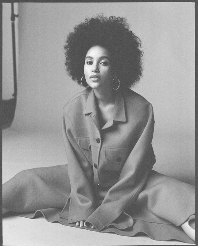 Portrait of a Black woman with afro, wearing an oversized suit, sitting on the floor in a studio with a neutral background.