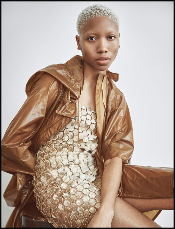 A young Black woman poses in a unique beige, short dress with a honey-brown hooded jacket in a studio setting, showcasing fashion and style.
