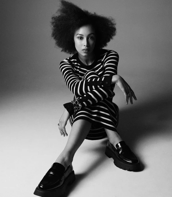 Black and white portrait of a woman with afro, wearing a striped dress and loafers, posing in a studio with soft, diffused lighting.