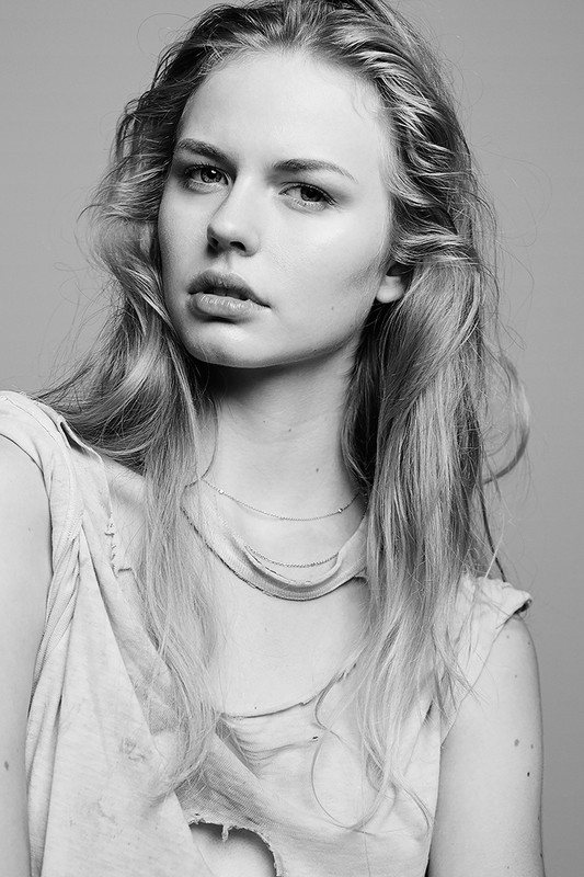 Black and white portrait of a young adult woman with long hair, wearing a ripped t-shirt, looking directly at the camera in a studio setting.