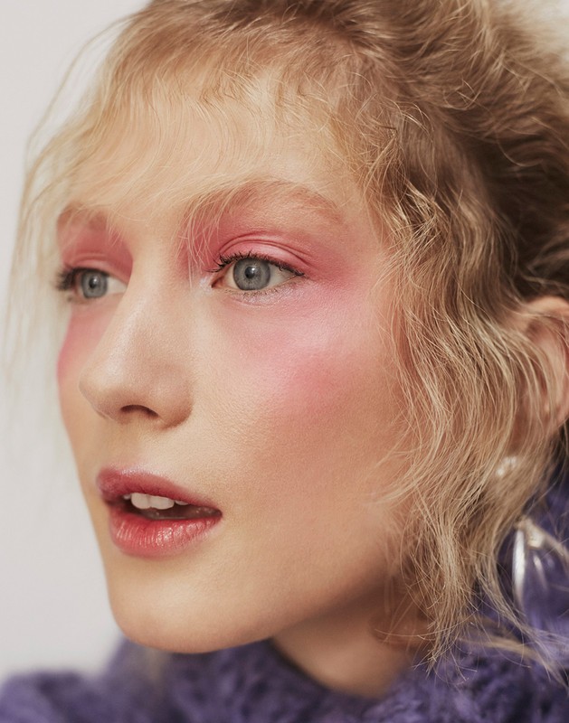 Close-up portrait of a fair-skinned young woman with pink eyeshadow, matching blush, and pink lipstick in front of a neutral background.