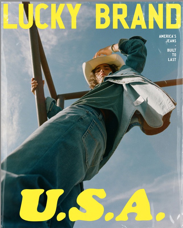 A denim-clad person in a cowboy hat poses with weathered metal against a blue sky in a Lucky Brand advertisement with U.S.A. text.