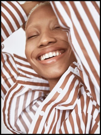 Close-up portrait of a smiling person with closed eyes wearing a brown and white striped button-down shirt with arms raised.