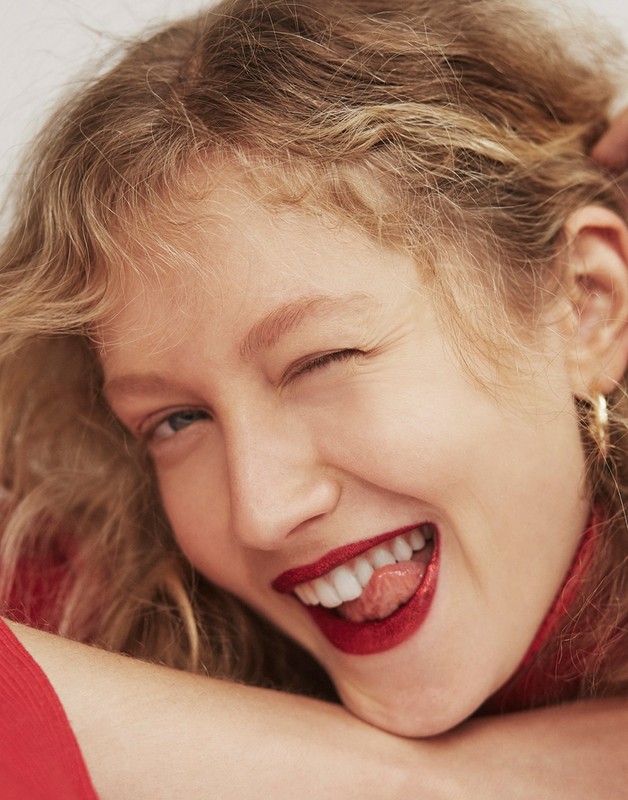 Close-up portrait of a blonde woman with curly hair, wearing red lipstick, sticking her tongue out, and winking in a beauty shot.