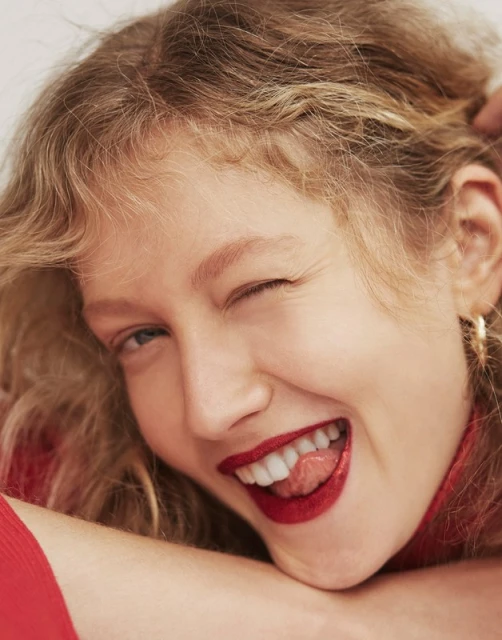Close-up portrait of a blonde woman with curly hair, wearing red lipstick, sticking her tongue out, and winking in a beauty shot.