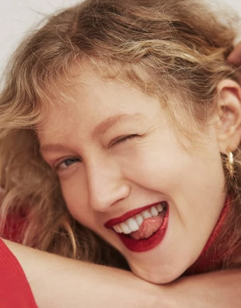 Close-up portrait of a blonde woman with curly hair, wearing red lipstick, sticking her tongue out, and winking in a beauty shot.
