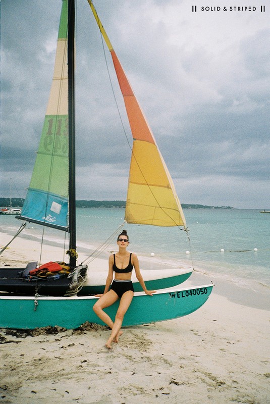 Adult female in a black bikini poses on a teal sailboat on a sandy beach, with the ocean and cloudy sky visible in the background.