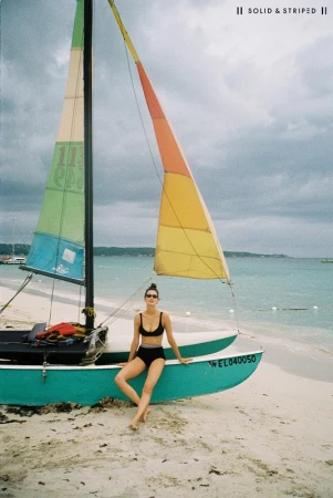 Woman in a black bikini sitting on the turquoise hull of a sailboat on a sandy beach under an overcast sky styled by Lindsay Grosswendt