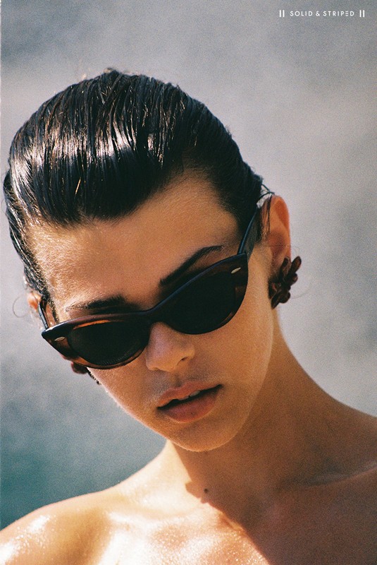 Close-up of a young adult woman wearing sunglasses and earrings, featuring slicked-back hair and a light complexion.