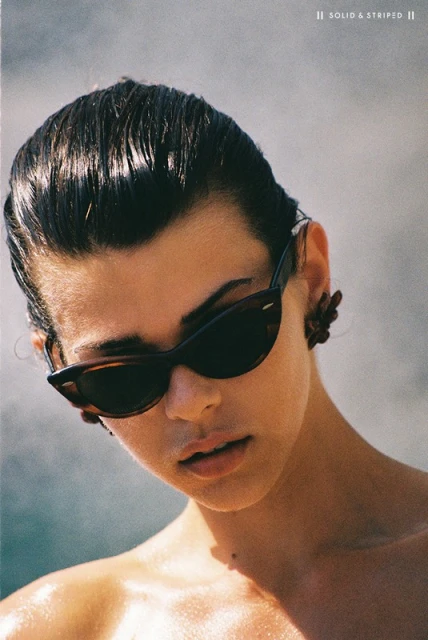 Close-up of a young adult woman wearing sunglasses and earrings, featuring slicked-back hair and a light complexion.