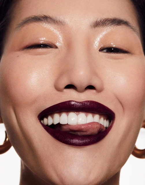 Close-up studio shot of an Asian adult female model with fair skin, dark burgundy lipstick, and gold hoop earrings with a bright smile.