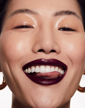 Close-up studio shot of an Asian adult female model with fair skin, dark burgundy lipstick, and gold hoop earrings with a bright smile.