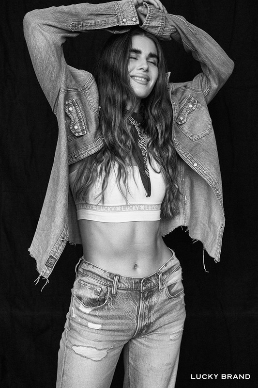 Black and white portrait of a woman with long wavy hair wearing an open denim shirt and distressed jeans, arms raised.