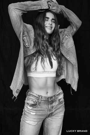 Black and white portrait of a woman with long wavy hair wearing an open denim shirt and distressed jeans, arms raised.