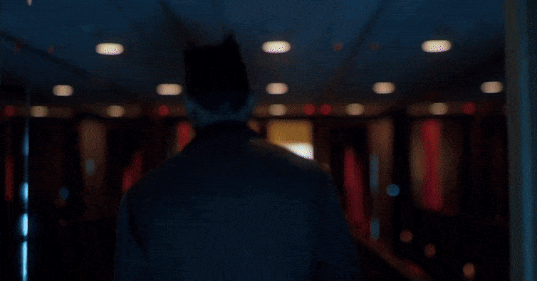 Back view of person walking down dimly lit interior hallway with paneled walls and ceiling lights. Interior architecture, dark mood.