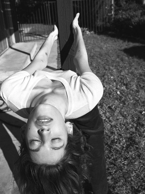 Black and white portrait of an adult woman with closed eyes holding a wooden post, captured outdoors on a sunny day.