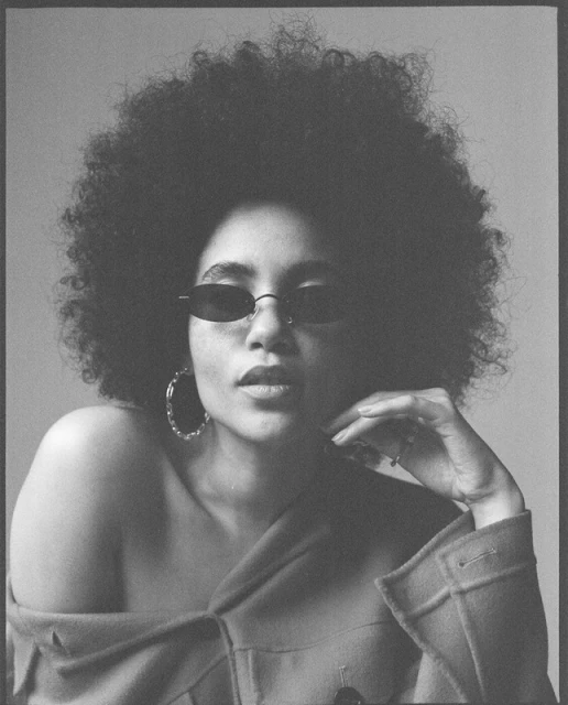 Black and white portrait of a stylish adult woman with an afro, wearing oval sunglasses and hoop earrings, posing in a photo studio.