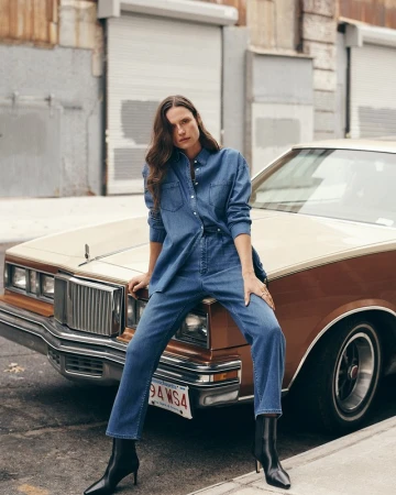 A woman in denim attire poses against a vintage car in an urban setting, styled with black heeled boots and a confident gaze.
