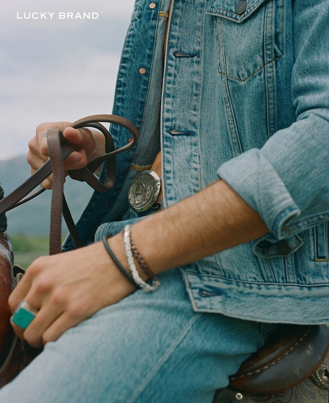 Man in denim jacket riding a horse outdoors. He wears bracelets and jeans. The image features the Lucky Brand logo in the top left.