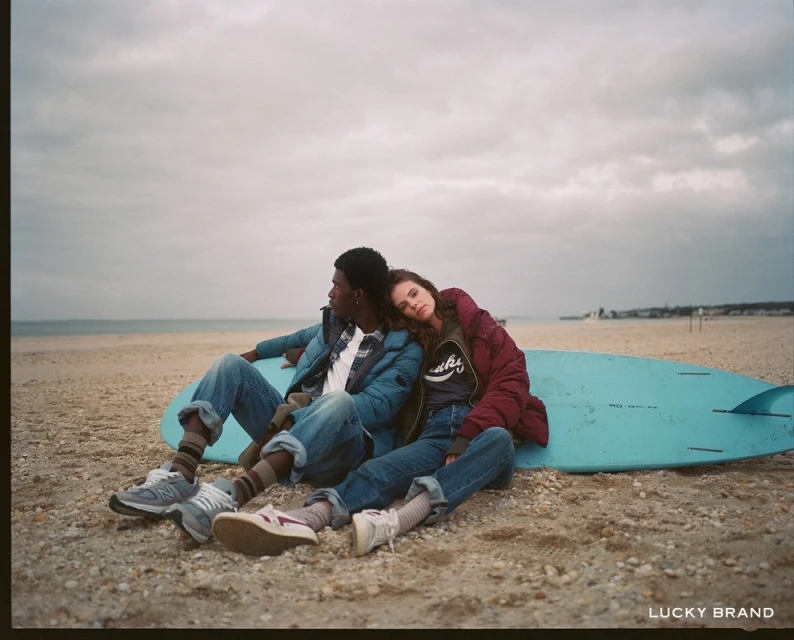 A couple relaxes on a beach with a surfboard under an overcast sky. A woman rests her head on the man's shoulder as they sit together.