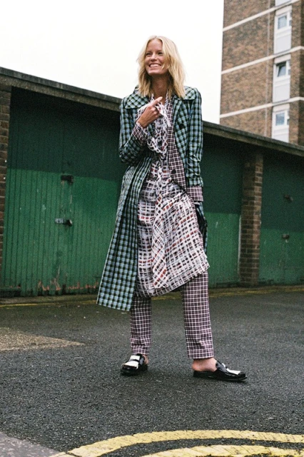 Smiling woman poses in an outdoor fashion shot wearing a green plaid coat, printed dress, and black shoes, with a building in the background.