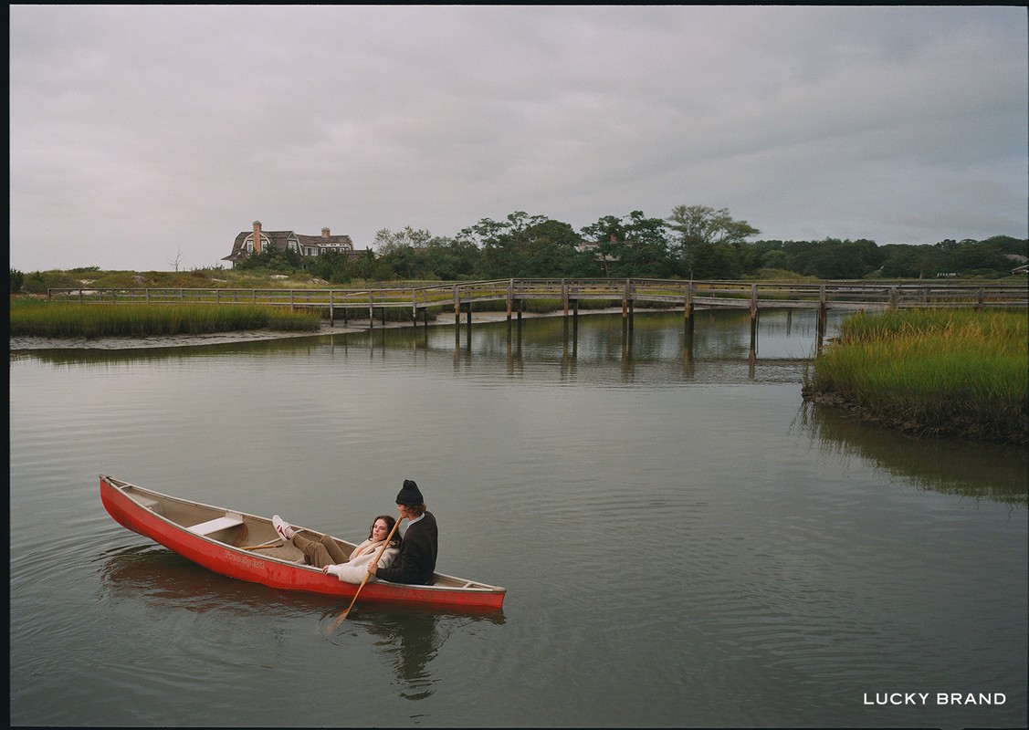 A couple paddles a red canoe on a calm river near a wooden bridge and grassy marsh under an overcast sky near a shoreline house.