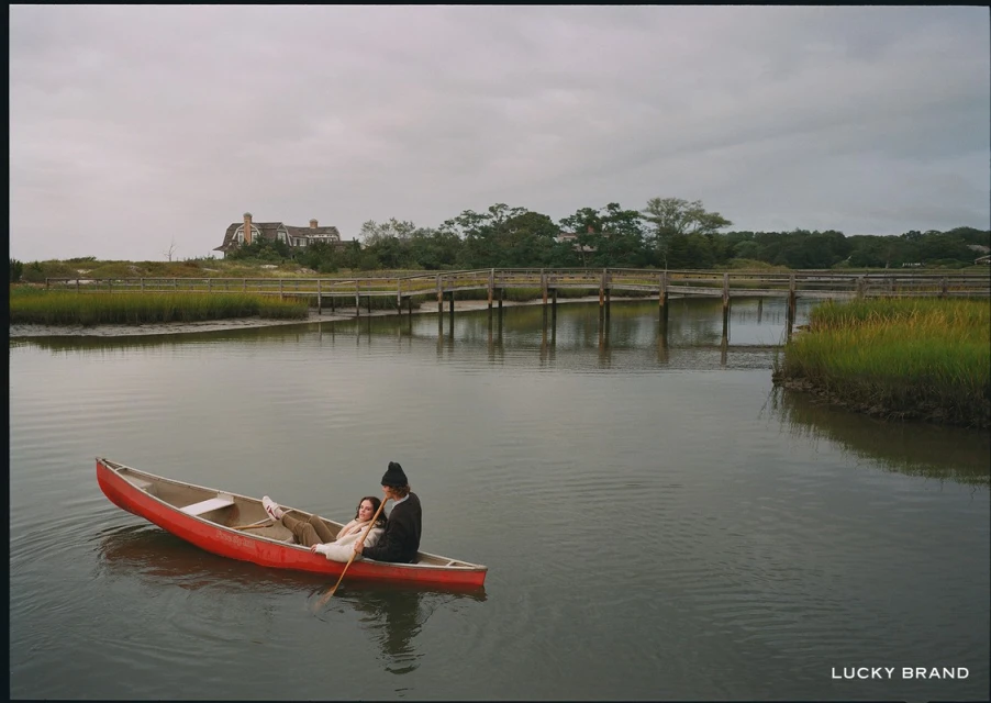 A couple paddles a red canoe on a calm river near a wooden bridge and grassy marsh under an overcast sky near a shoreline house.