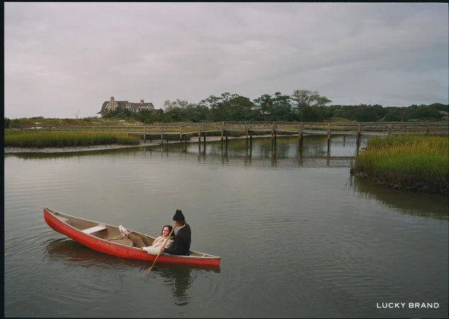 A couple paddles a red canoe on a calm river near a wooden bridge and grassy marsh under an overcast sky near a shoreline house.