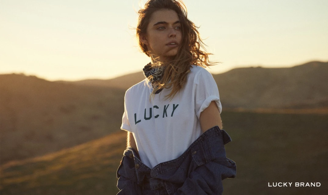 Portrait of a young adult woman wearing a white 'Lucky' t-shirt and denim jacket with a sunset and hilly landscape background.