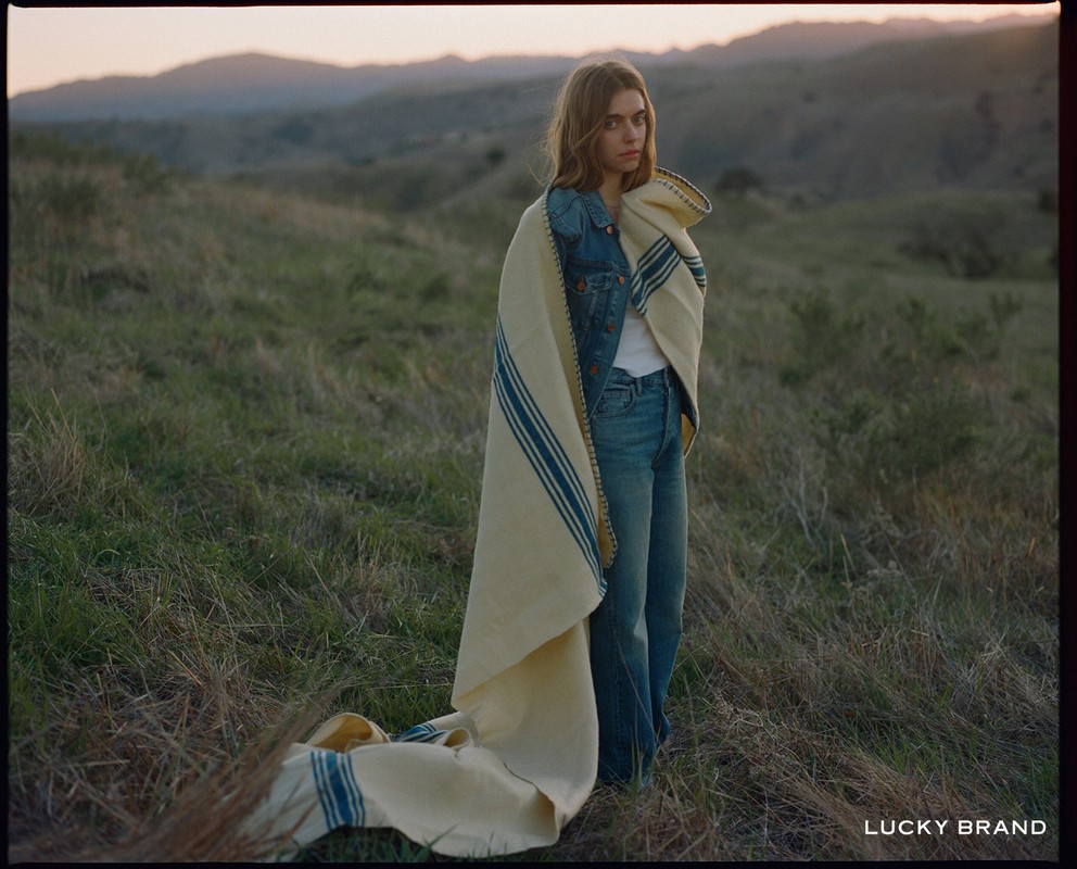 A young woman stands in a field wrapped in a striped blanket, wearing a denim jacket and jeans, against a backdrop of grassy hills.