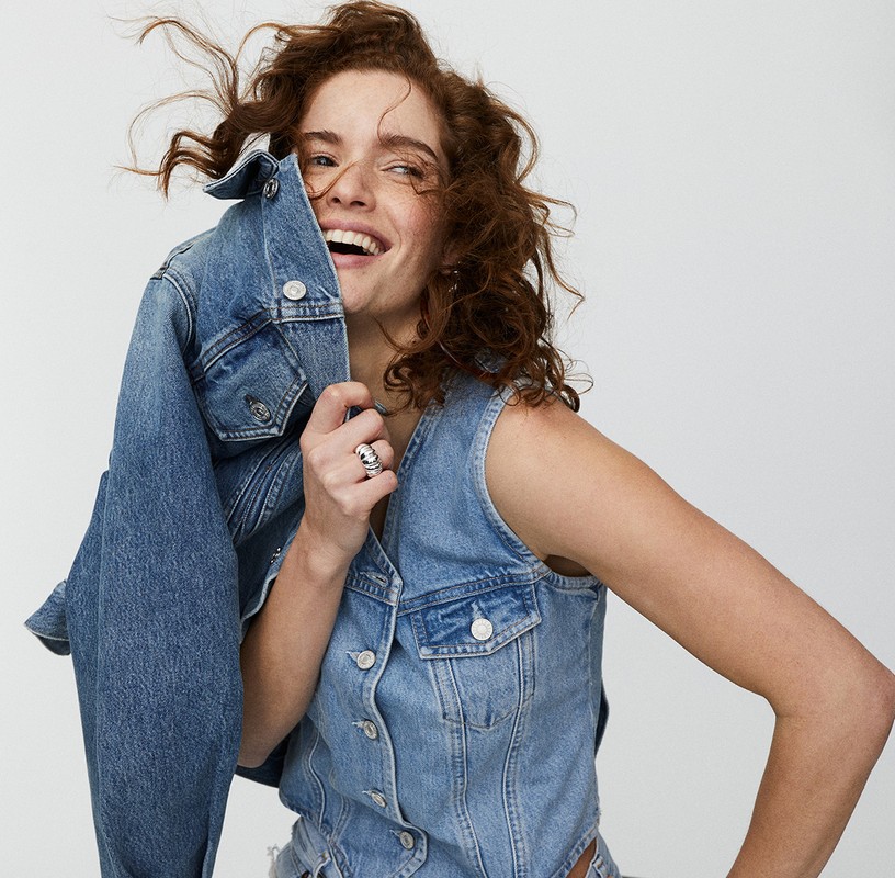 Smiling woman with red hair wearing a denim vest poses in front of a neutral background, holding a denim jacket in her hand.