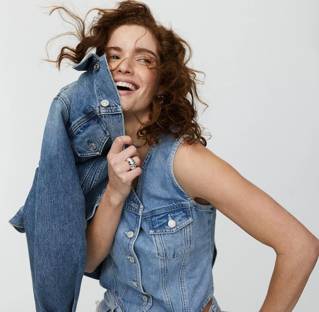 Smiling woman with red hair wearing a denim vest poses in front of a neutral background, holding a denim jacket in her hand.