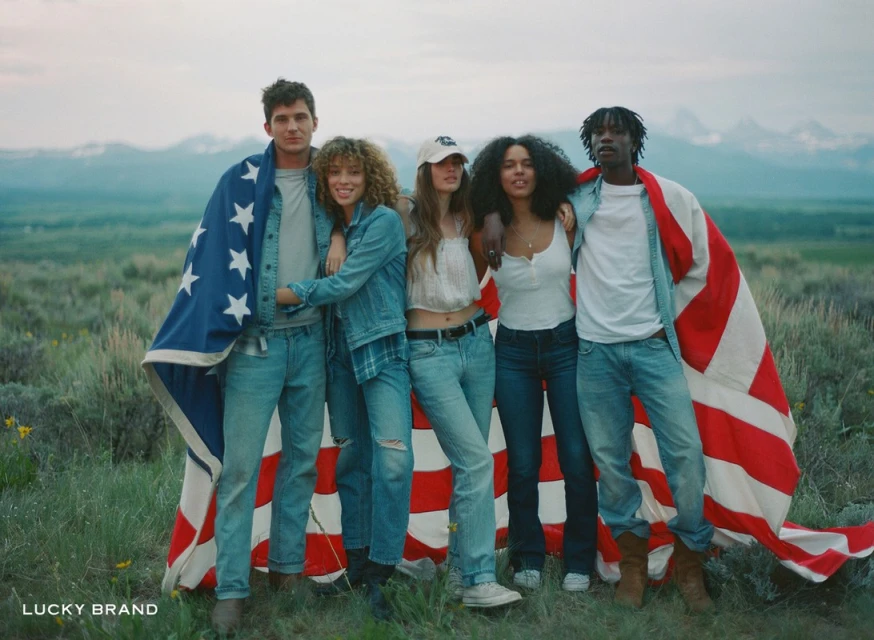 A group of five young adult models in denim and casual tops pose in a grassy field holding a large American flag with mountains in the background.