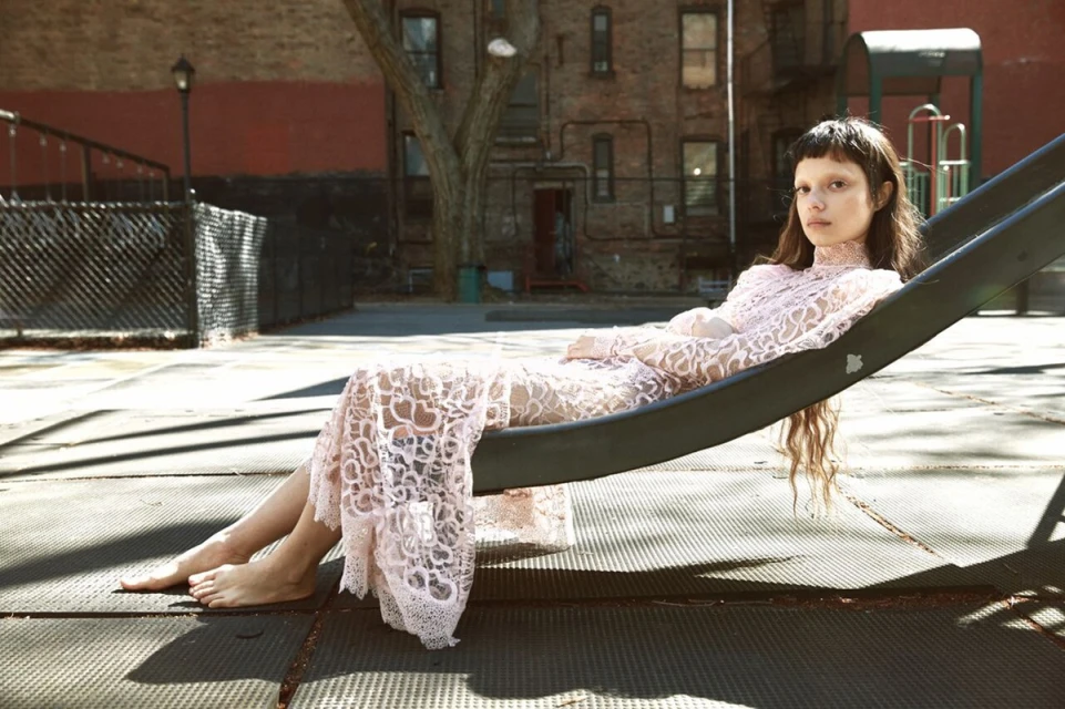 A woman in a light pink lace dress is lying on a playground slide on a sunny day in an urban environment.
