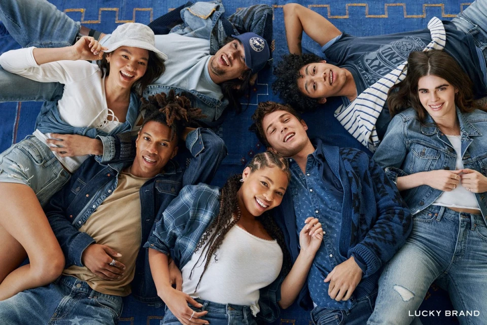 Overhead shot of seven smiling adults laying on a blue patterned rug, wearing a variety of light and dark denim clothing and posing for the camera.