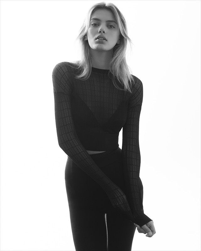 Blonde woman in a dark, long-sleeved sheer top and dark leggings poses in front of a bright white background in a studio portrait.