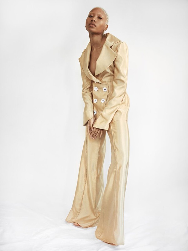 Model with short platinum blonde hair wearing a gold satin pant suit with large white buttons against a plain white background
