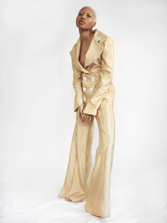 Model with short platinum blonde hair wearing a gold satin pant suit with large white buttons against a plain white background