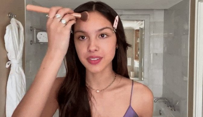 Olivia Rodrigo applying makeup with a brush in a hotel bathroom, styled by Lindsay Grosswendt in Los Angeles.