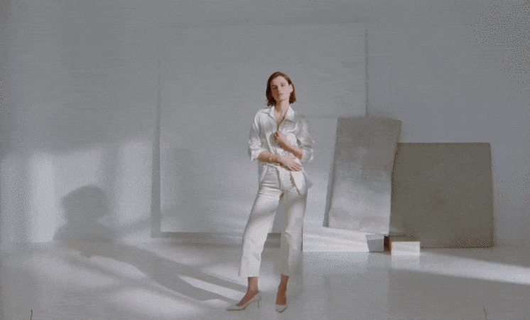 A woman poses in a light, all-white pantsuit with an unbuttoned blouse and matching heels in a bright, minimalist photo studio setting.