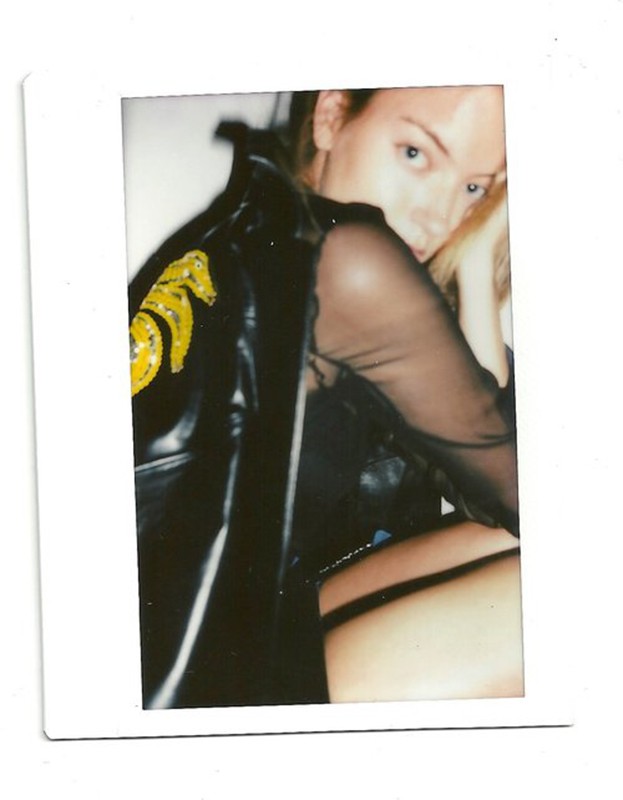 Blonde-haired woman in black sheer top and leather jacket with embellished seahorse design on the back, in a polaroid-style frame.