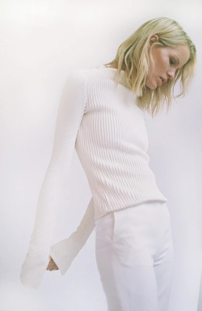 A solitary blonde female poses indoors, wearing a white, long-sleeved ribbed top and matching pants against a stark white background.