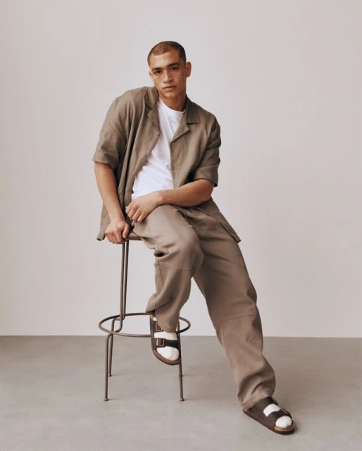 Man in a beige linen outfit and sandals sitting on a metal stool in a studio with a neutral background in a fashion setting.