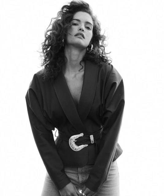 Black and white portrait of a woman with curly hair wearing a blazer with a decorative belt and denim jeans against a white background.
