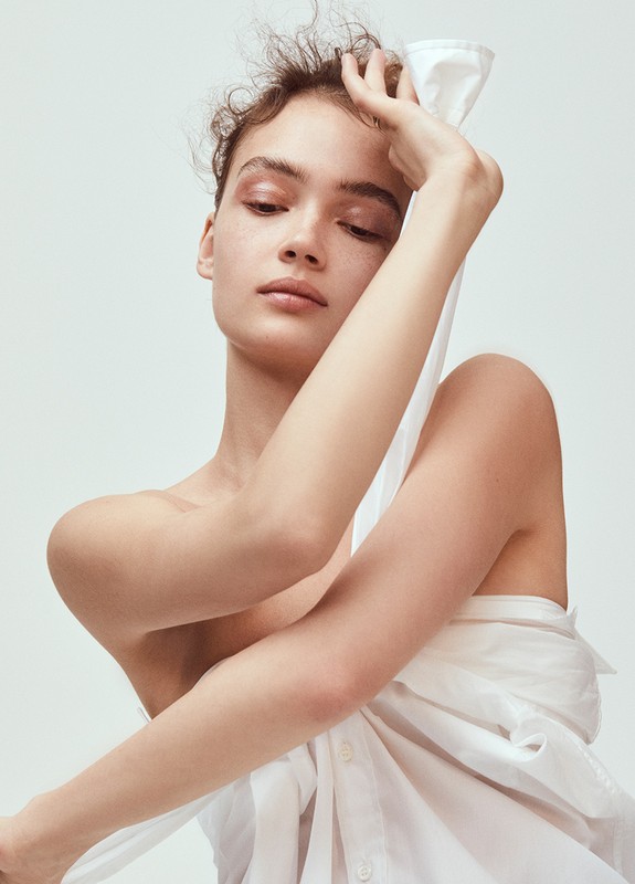 A young adult female with curly brown hair, adorned in subtle makeup, poses in a studio. She is draped in a white collared shirt.