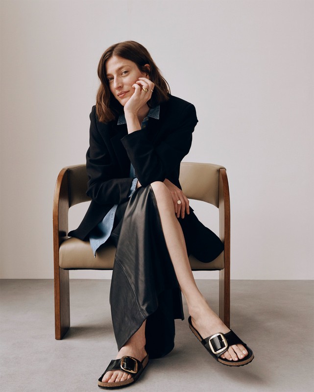 Brunette woman poses seated in chair, wearing black blazer, denim shirt, long black skirt, and black sandals with gold buckles.