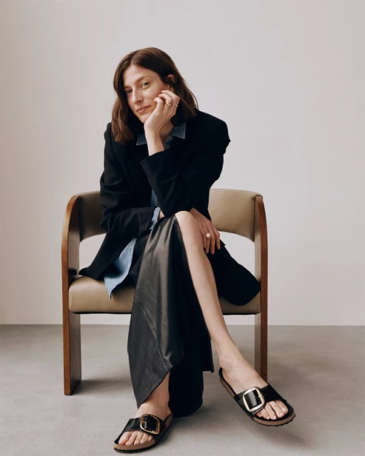 Brunette woman poses seated in chair, wearing black blazer, denim shirt, long black skirt, and black sandals with gold buckles.