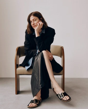 Brunette woman poses seated in chair, wearing black blazer, denim shirt, long black skirt, and black sandals with gold buckles.