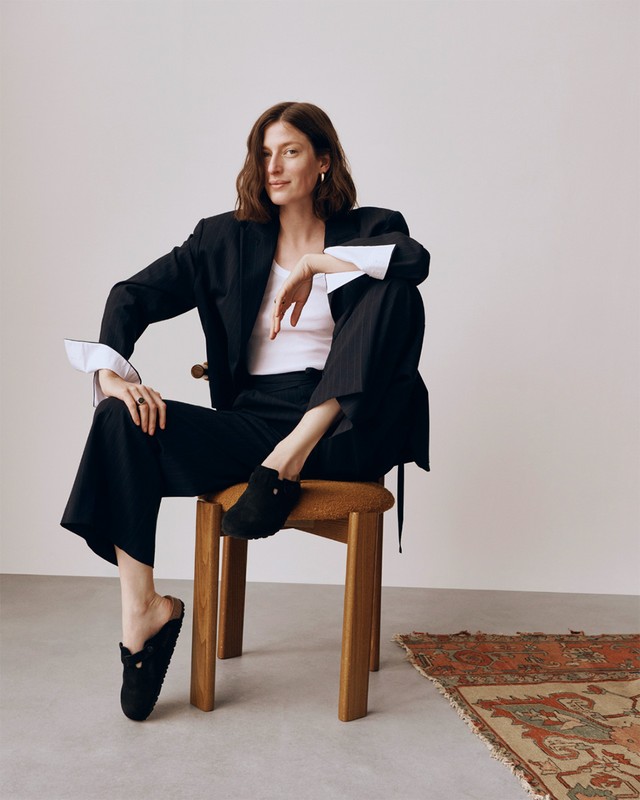 A woman sits on a wooden stool in a black blazer, white top, and wide-legged trousers, posing in an indoor studio setting.
