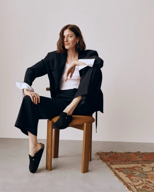 A woman sits on a wooden stool in a black blazer, white top, and wide-legged trousers, posing in an indoor studio setting.